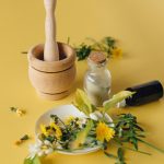 Ingrédients pour un remède naturel à base de plantes avec un mortier, un piédestal et une bouteille sur une surface jaune.