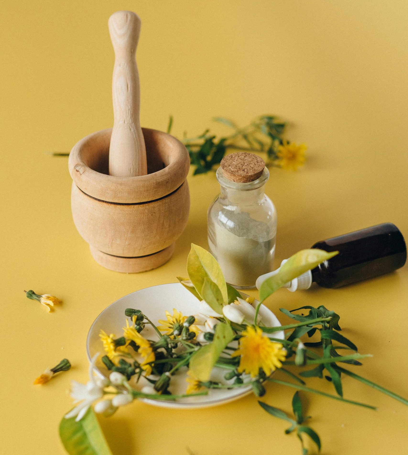 Ingrédients pour un remède naturel à base de plantes avec un mortier, un piédestal et une bouteille sur une surface jaune.